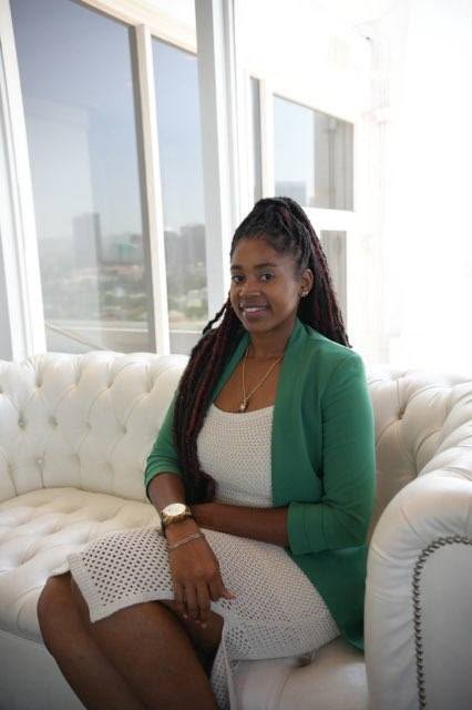 Lovett Love seated on a white tufted sofa in a green blazer, with the city skyline through the windows behind her
